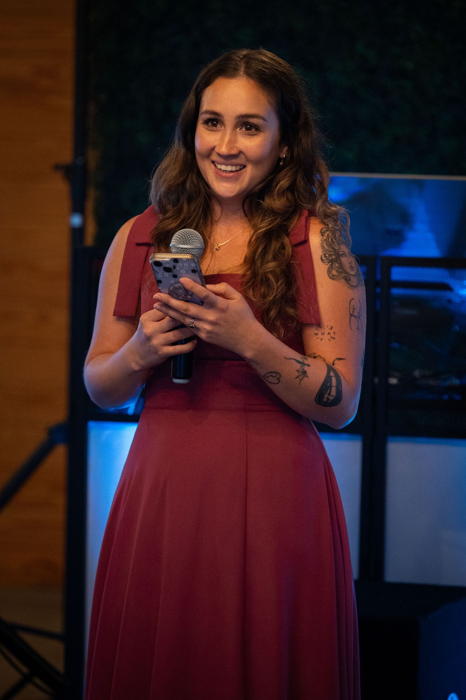 A smiling woman in a red dress holds a microphone while speaking at an indoor event with blue lighting.