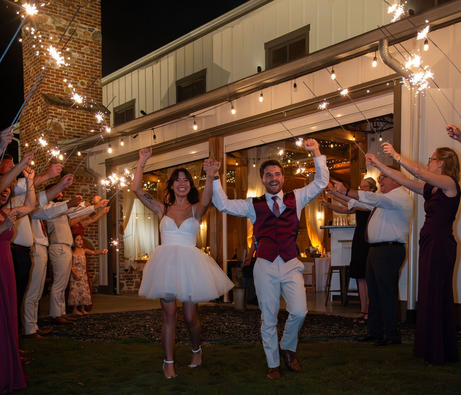 Bride and groom exit wedding reception through sparkler tunnel with cheering guests at night.