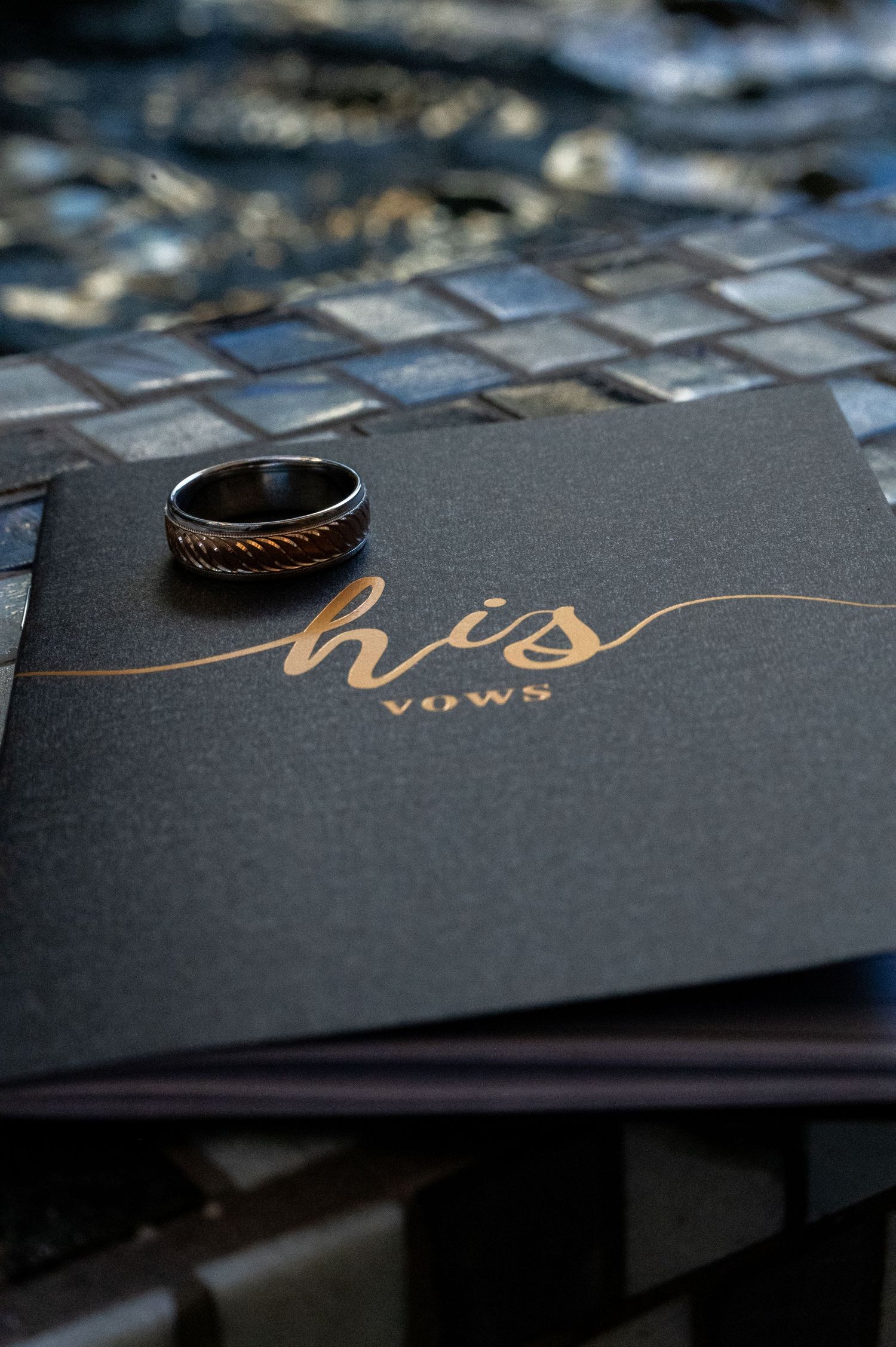 Black wedding ring resting on a dark vow book with gold script reading his vows on a mosaic tile surface.