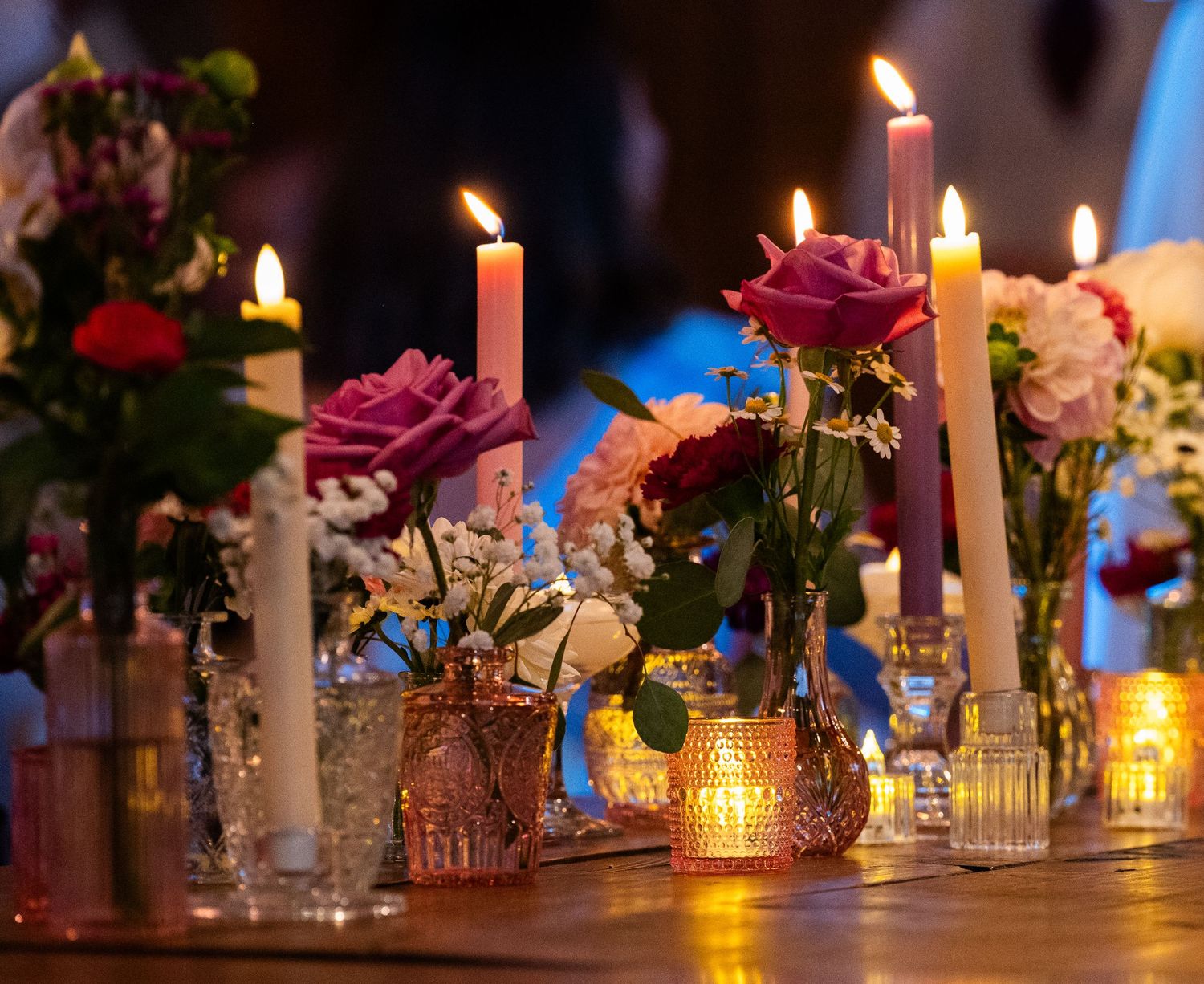 Elegant wedding table centerpiece with lit candles and pink roses in glass vases on a wooden table.