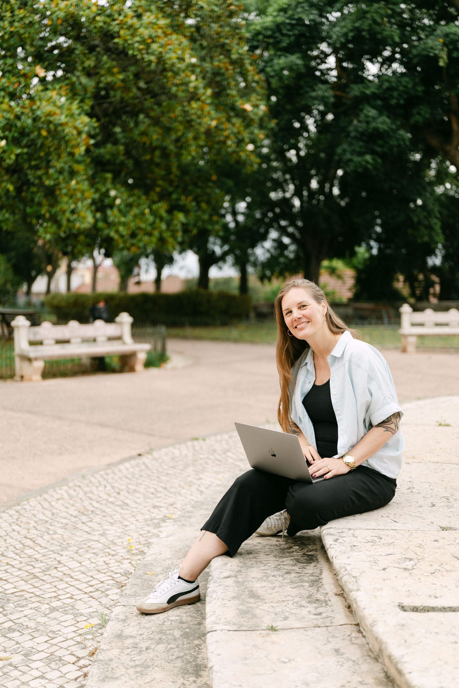 Smiling woman sitting outdoors on stone steps with laptop, surrounded by green trees in a park setting.