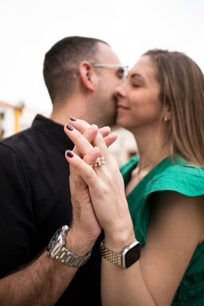 Couple sharing a kiss while holding hands, showcasing an engagement ring, man in black shirt, woman in green dress.