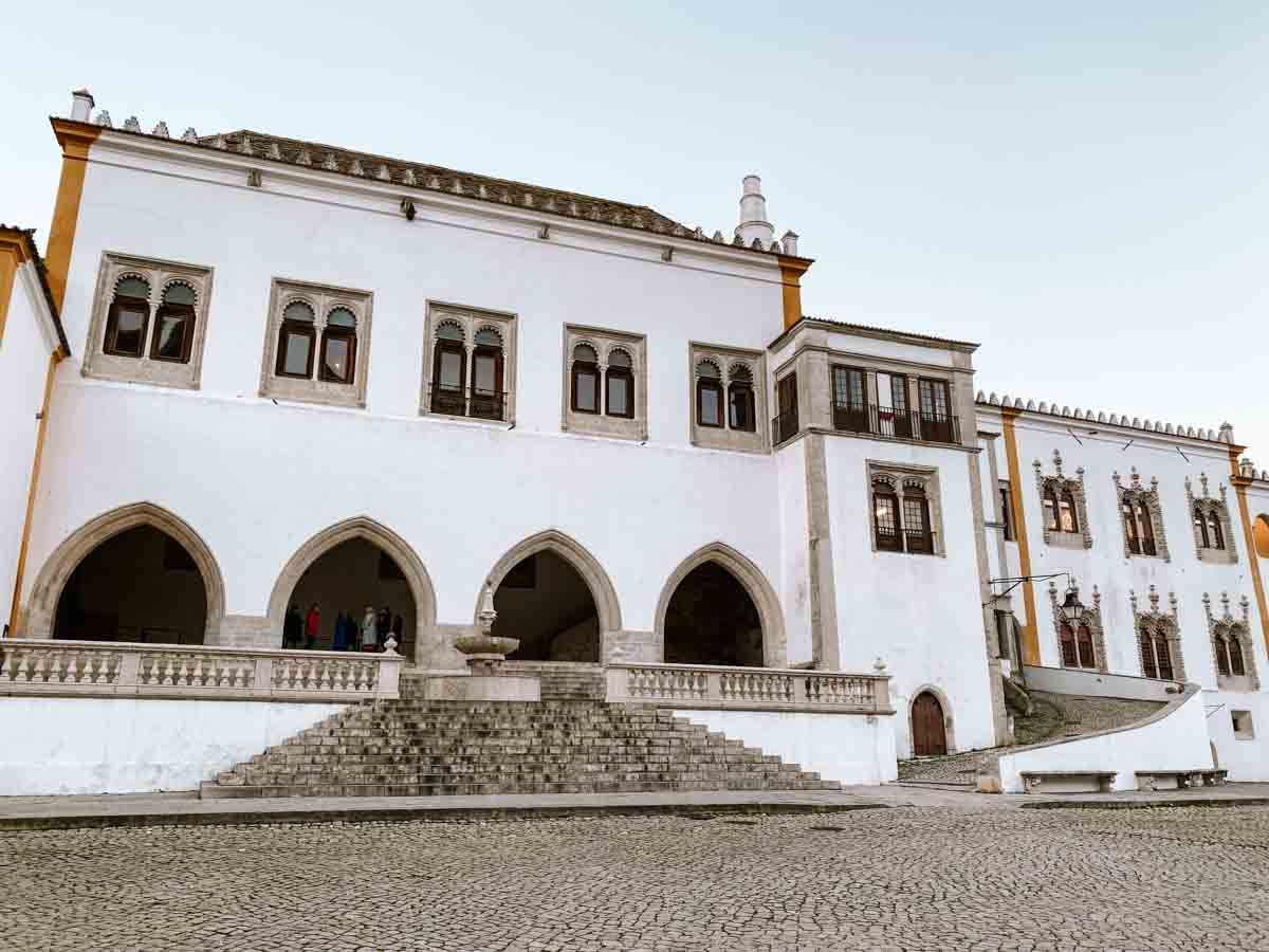 Historic white Portuguese palace with arched portico, ornate windows, and grand stone staircase in a sunlit courtyard.