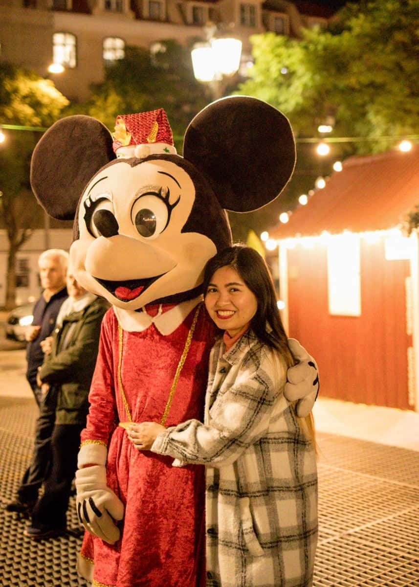 A woman poses with a Minnie Mouse character in a red outfit at an outdoor evening event with festive lights.