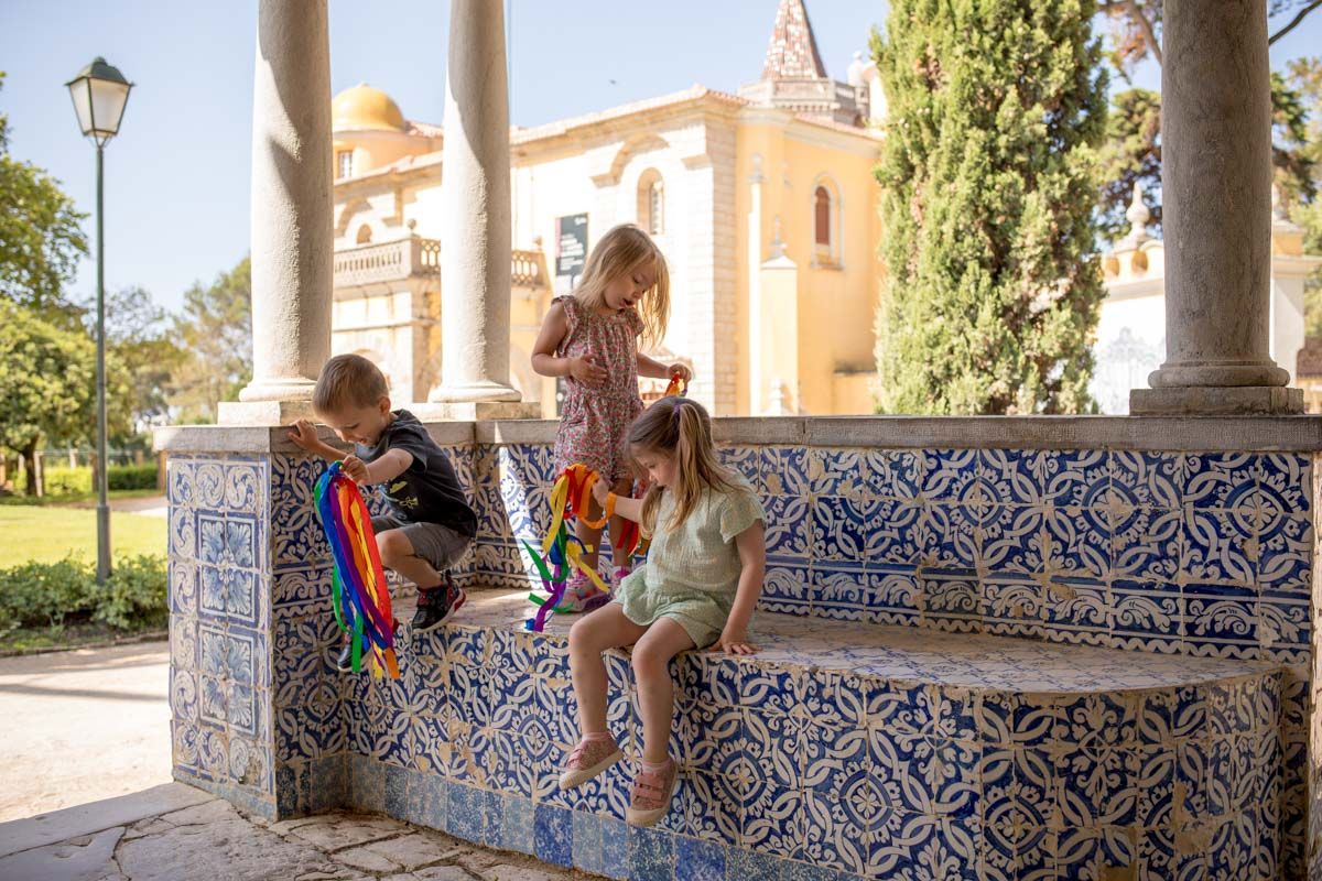 Family photoshoot Cascais with three young children playing on a blue tiled bench beneath stone columns in a sunny garden.