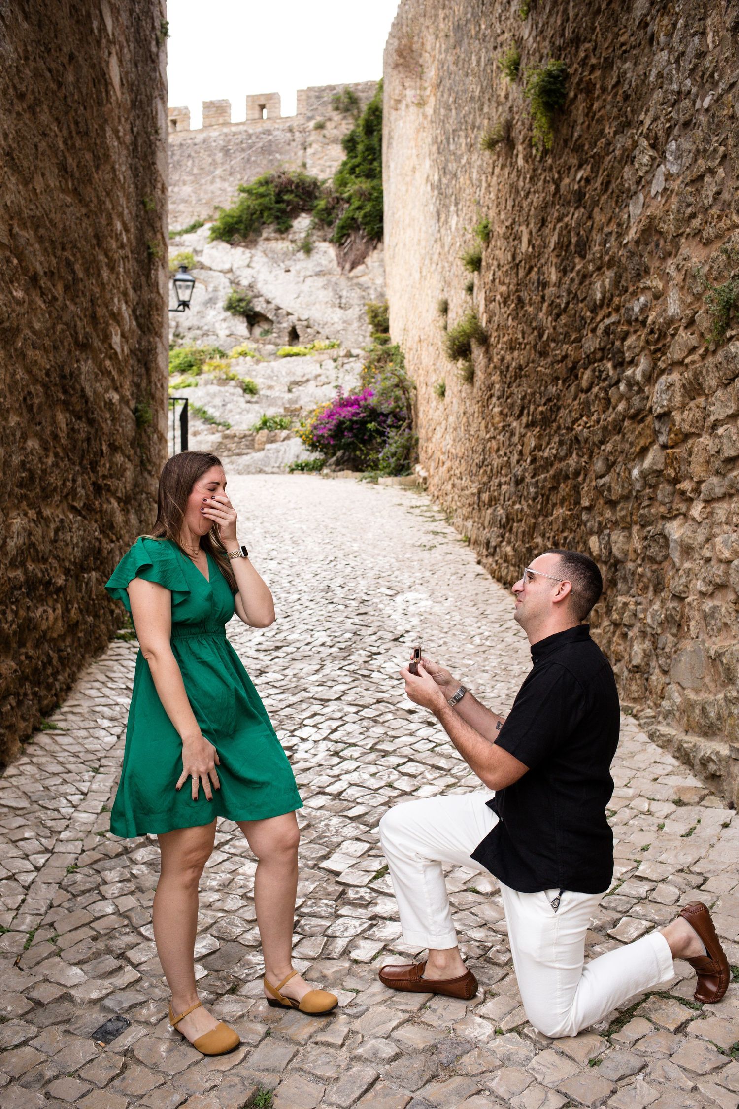 Man proposes on one knee to surprised woman in green dress on a cobblestone alley beside ancient stone walls.