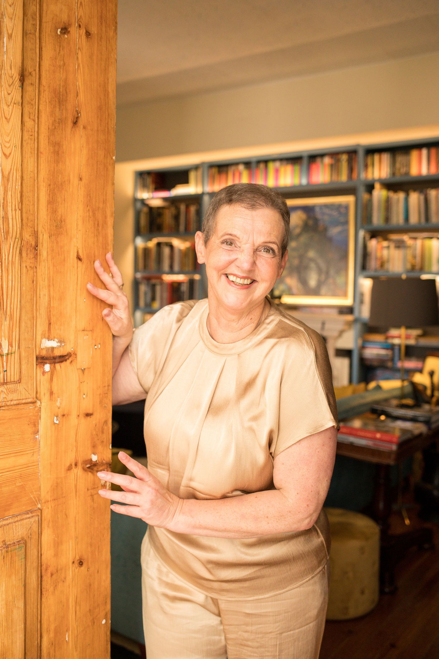 Smiling woman in a satin champagne blouse stands beside a rustic wooden door in a cozy home filled with bookshelves and warm lighting.