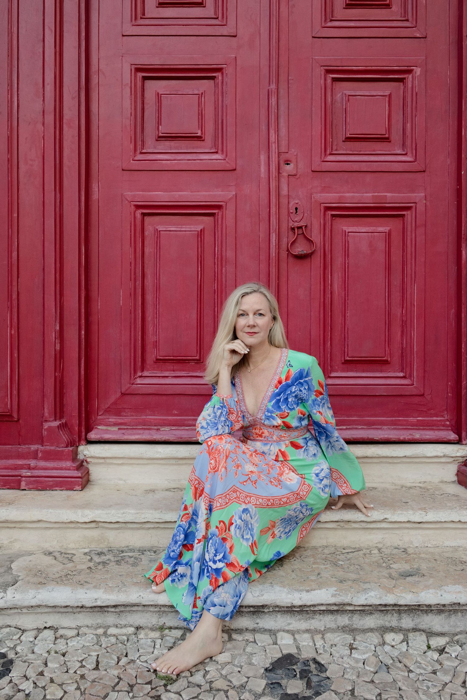 Blonde woman in a colorful floral maxi dress sits barefoot on stone steps in front of a large red paneled wooden door.