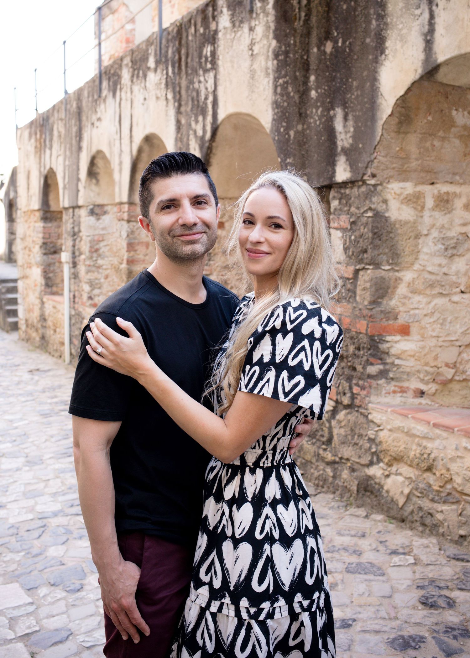 Couple embracing and smiling in front of a historic stone wall on a cobblestone street.