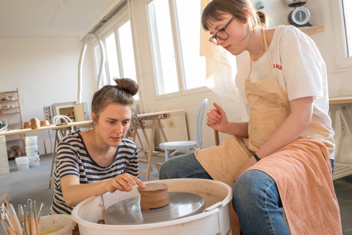 Two women in aprons work at a pottery wheel in a bright ceramics studio, shaping wet clay during a hands-on pottery class.
