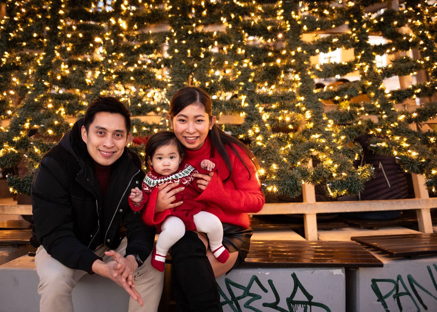 Family of three posing in front of a glowing Christmas tree with warm lights during the holiday season.