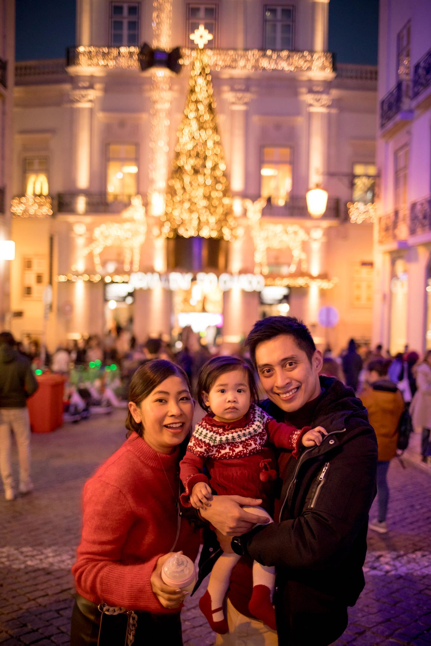 Asian family with toddler posing before a glowing Christmas tree and festive lights at a European plaza at night.