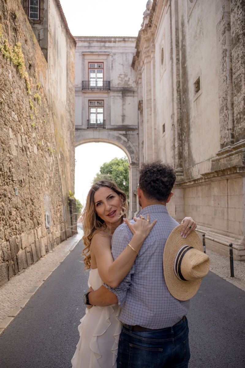 Couple embracing on a cobblestone street beneath a historic stone arch in a European city.