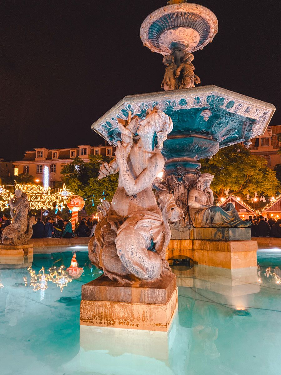 Illuminated baroque fountain with mythological figures at night, glowing blue water, festive lights in background.