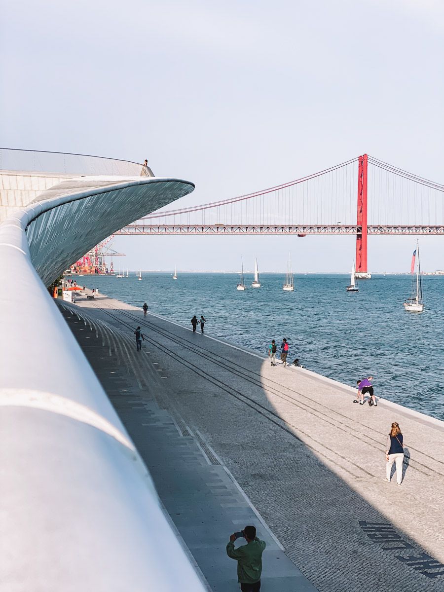 Lisbon waterfront promenade beside MAAT museum with the red 25 de Abril suspension bridge over the Tagus River.