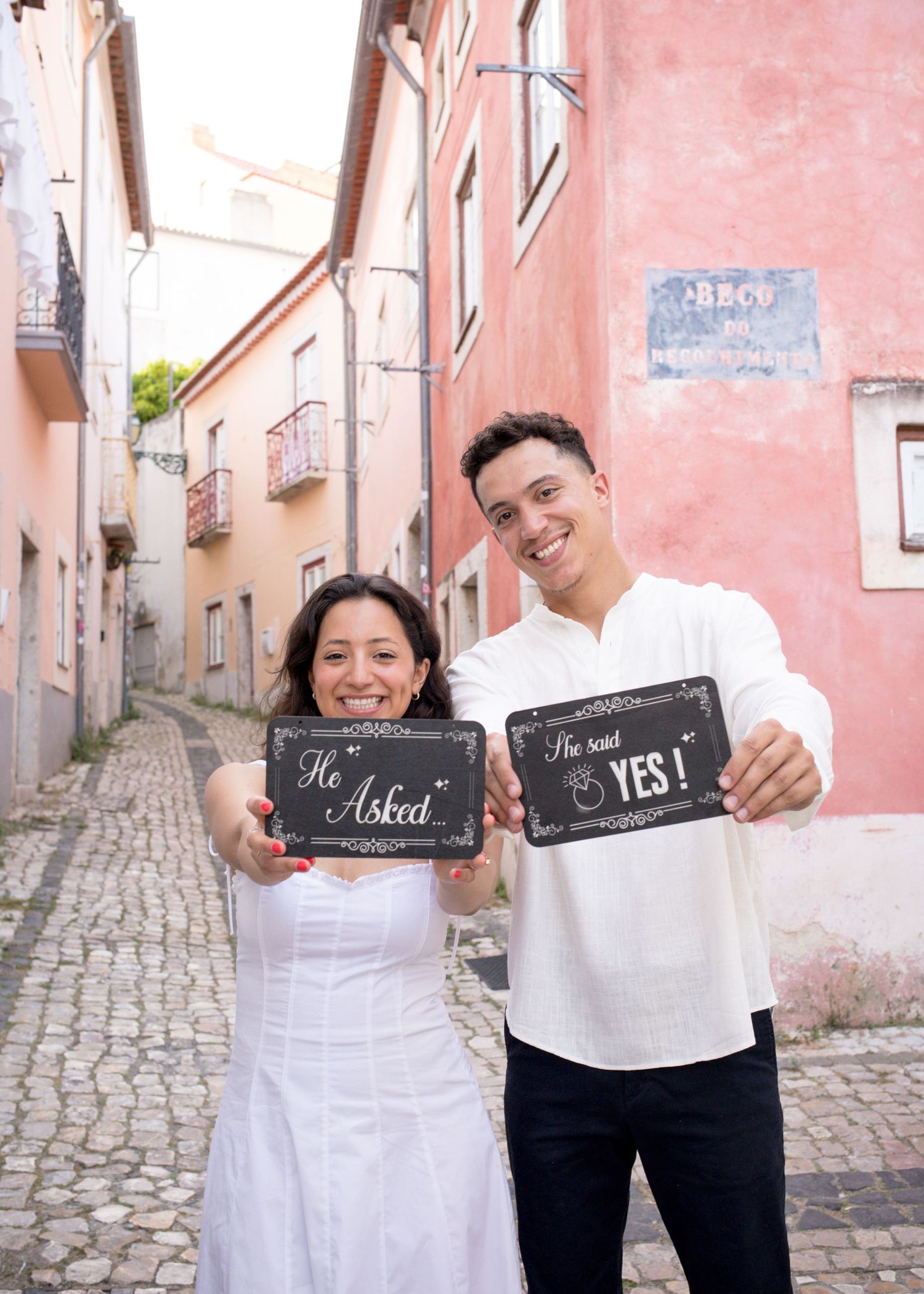 Couple holding He Asked and She Said Yes chalkboard signs on a cobblestone street in Lisbon, Portugal.