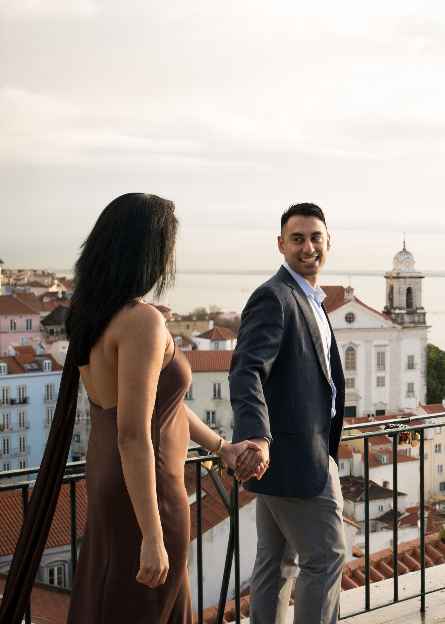 Couple holding hands on a scenic Lisbon balcony, man in navy suit smiling, city rooftops and church visible behind.