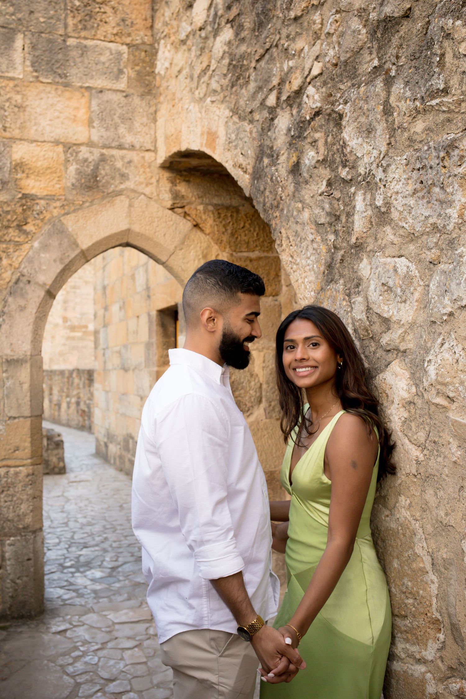 Couple holding hands and smiling against ancient stone arch wall, woman in green dress, man in white shirt.