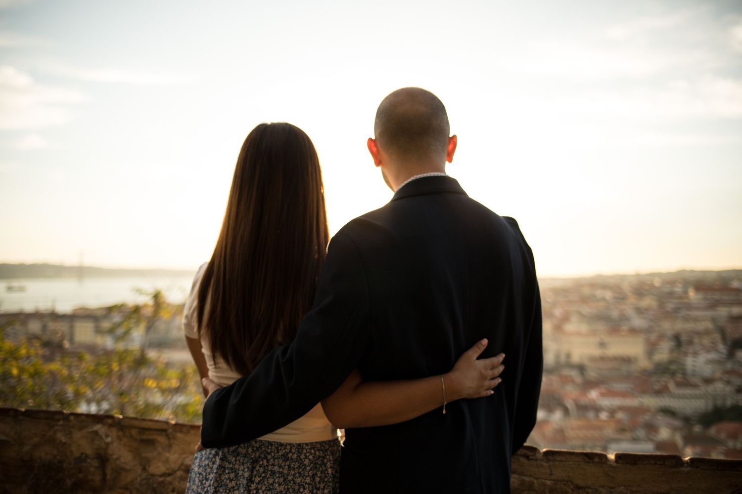 Couple embracing while overlooking a scenic city skyline at sunset from a hilltop viewpoint.