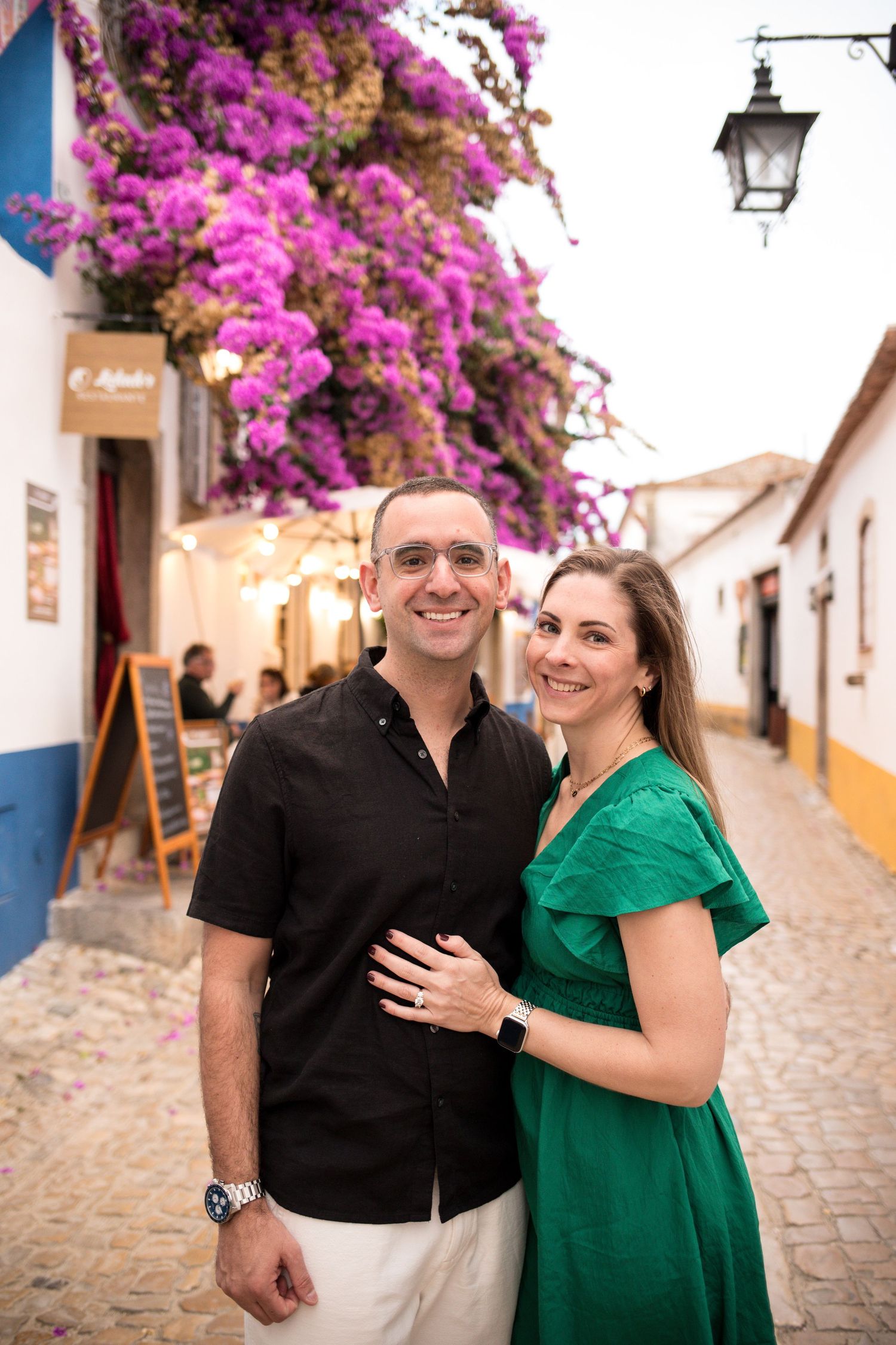 Proposal in Óbidos portrait of a smiling couple on a cobbled street beneath bright pink bougainvillea after getting engaged.
