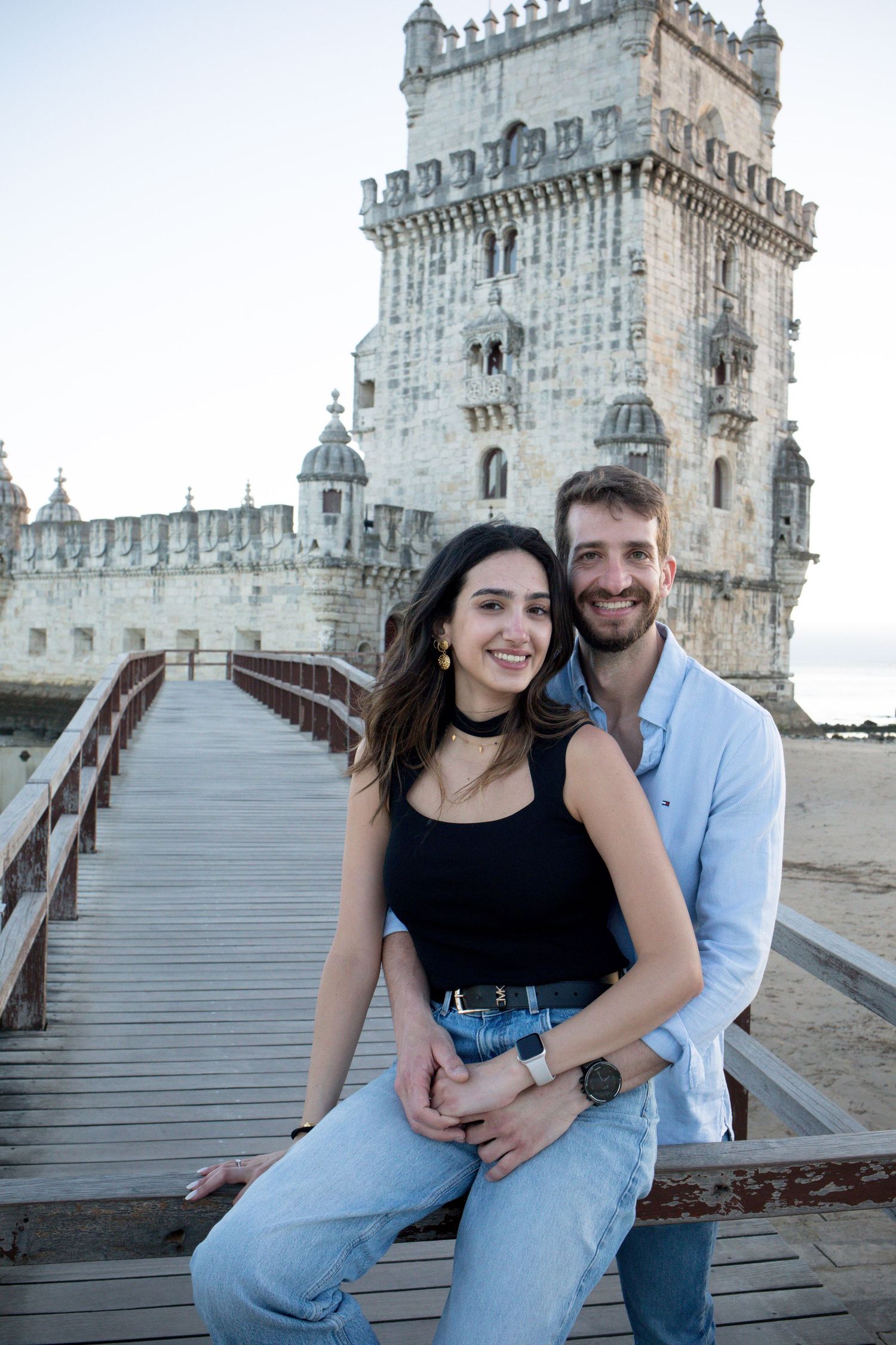 Smiling couple sits together on wooden bridge with iconic Belem Tower in Lisbon behind them.
