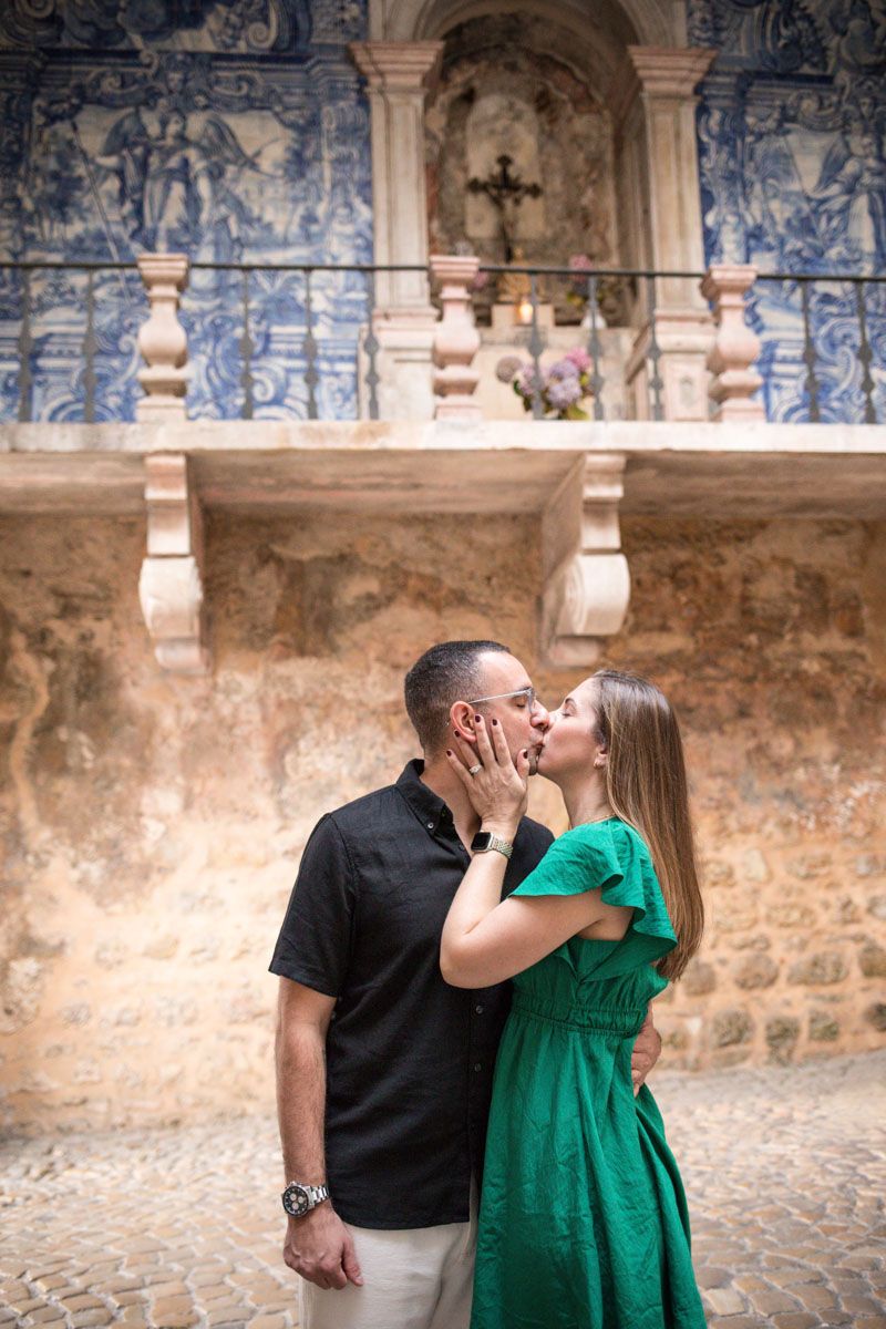 Couple sharing a romantic kiss in front of an ornate Portuguese azulejo tile wall and stone balcony.