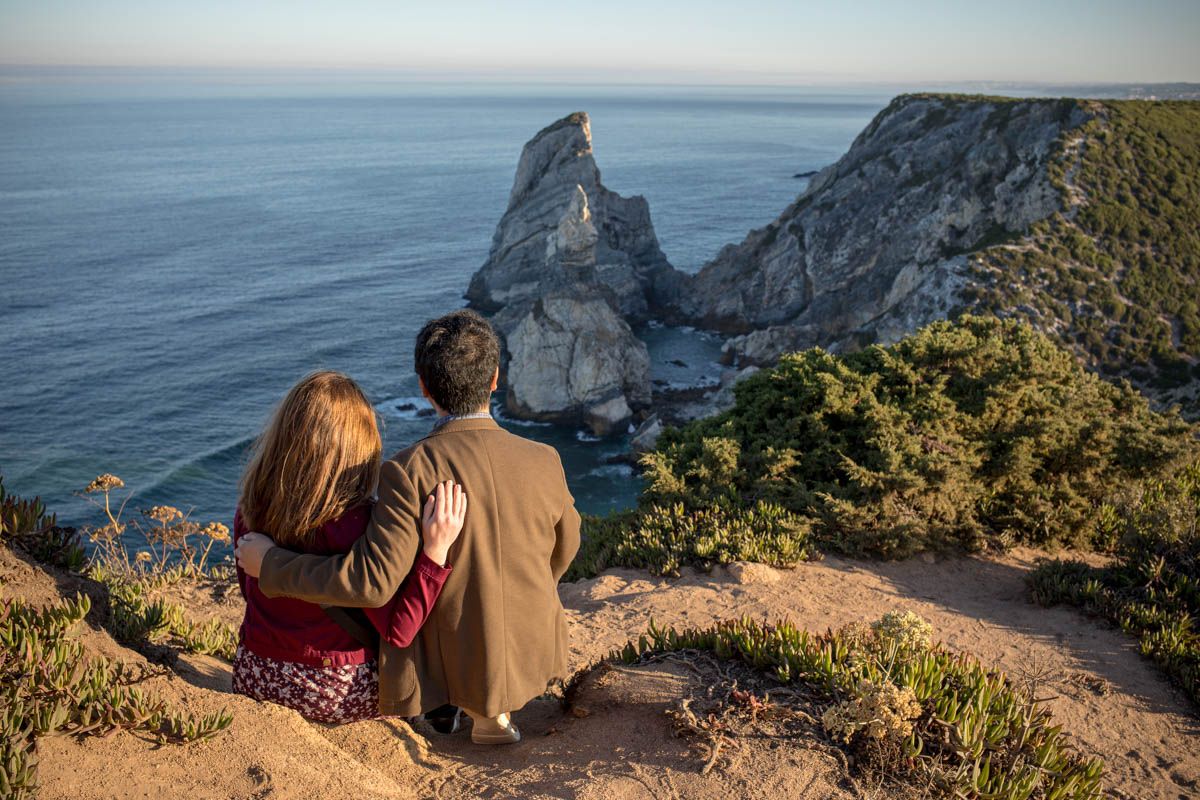 Couple sitting together on a cliff edge overlooking dramatic ocean rock formations at golden hour.