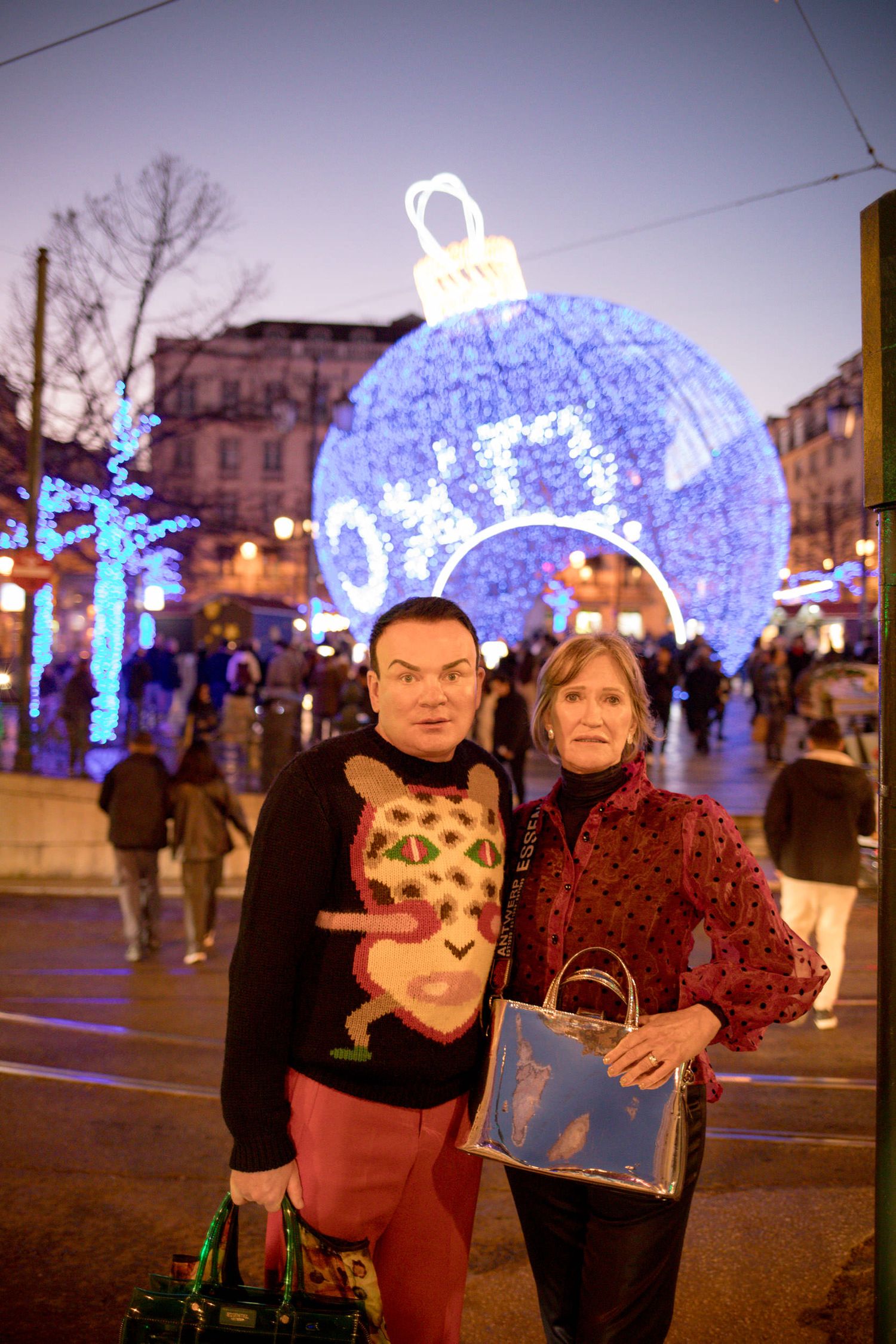 Couple posing in front of a giant blue Christmas ornament light display on a festive city street at dusk.