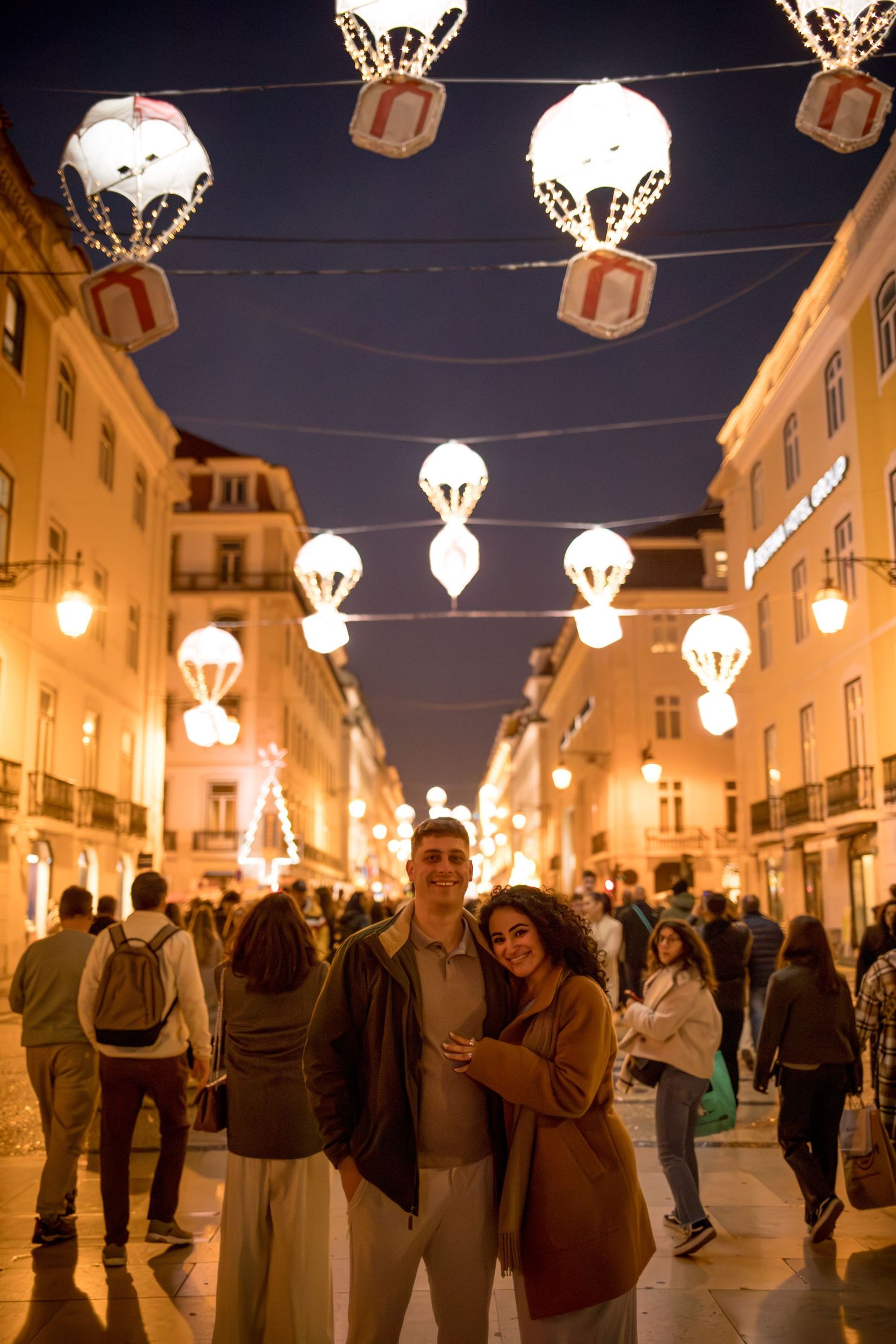 Couple embracing on a festive night street adorned with glowing parachute lights and holiday decorations in a busy city.