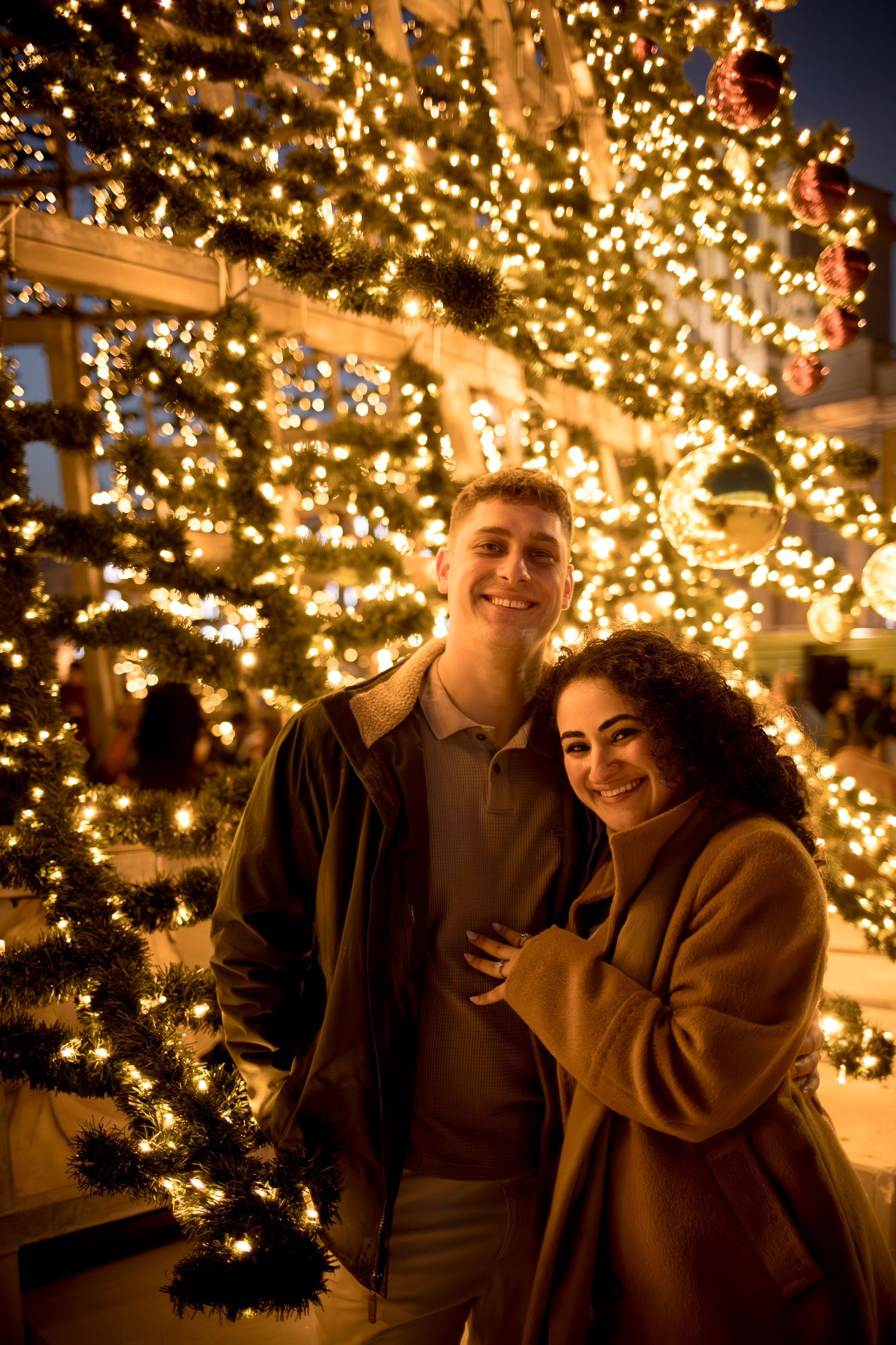 Smiling couple posing in front of a beautifully lit Christmas tree with golden lights at night.