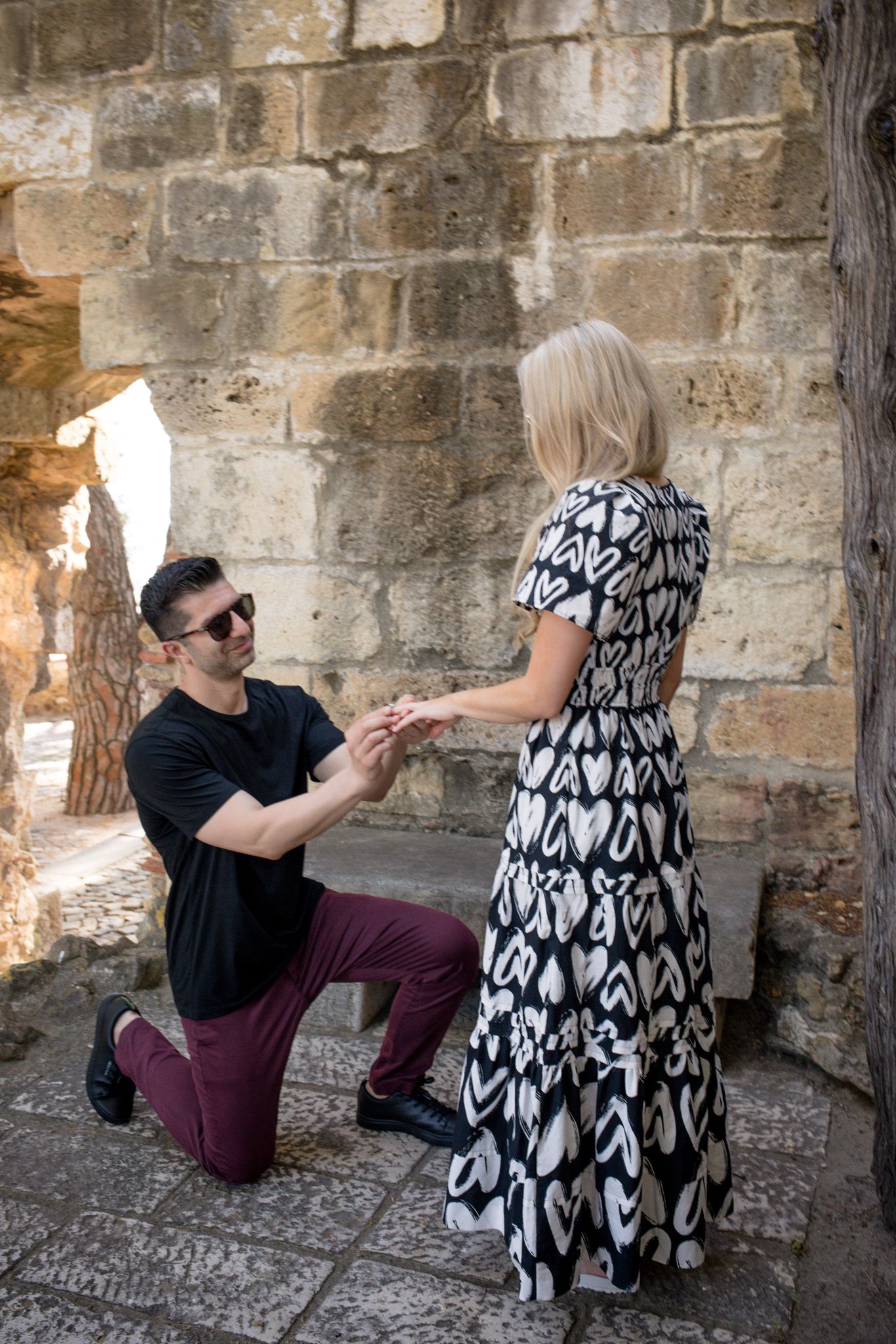 Man proposes on one knee to woman in black and white heart dress against ancient stone wall.