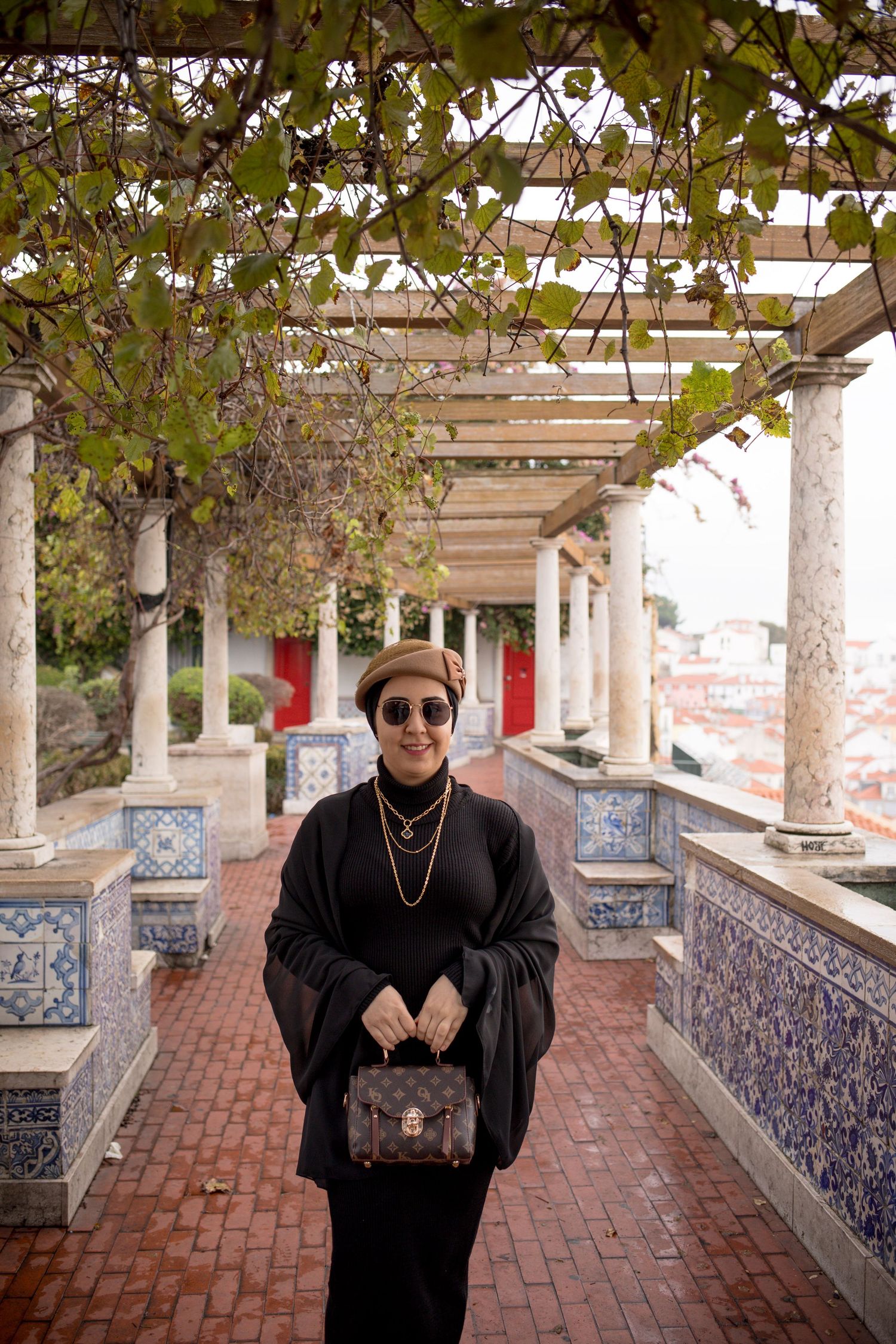 Woman in black outfit with beret and handbag posing under pergola with tiled walls.