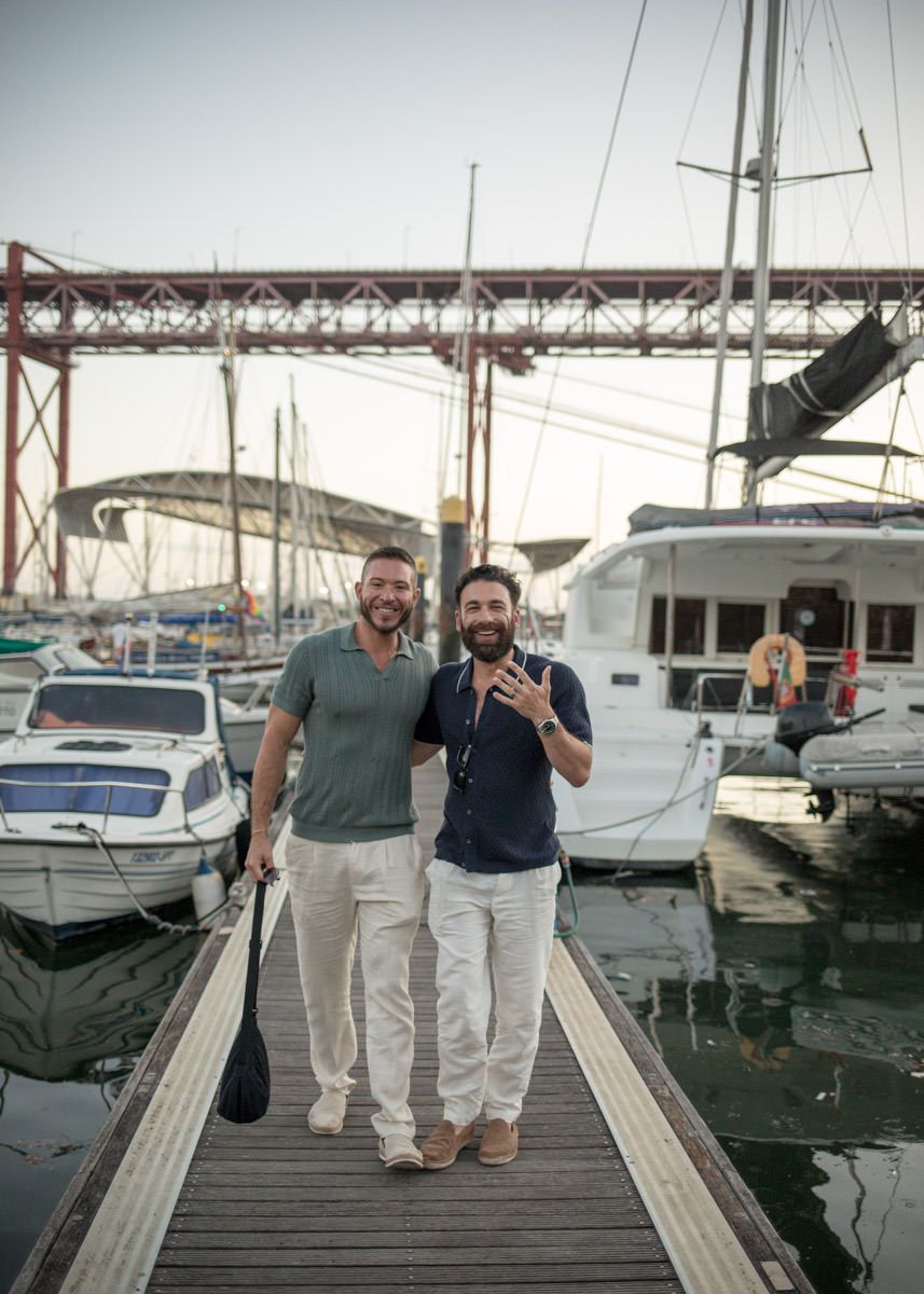 Two smiling men walk along a marina dock with sailboats and a red suspension bridge in the background.
