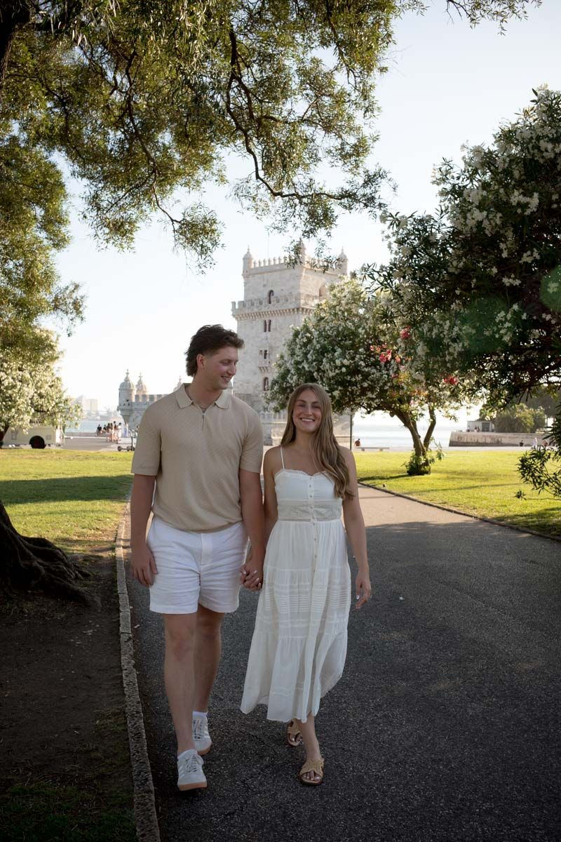 A couple holds hands walking near Belem Tower in Lisbon, Portugal, dressed in summer outfits.