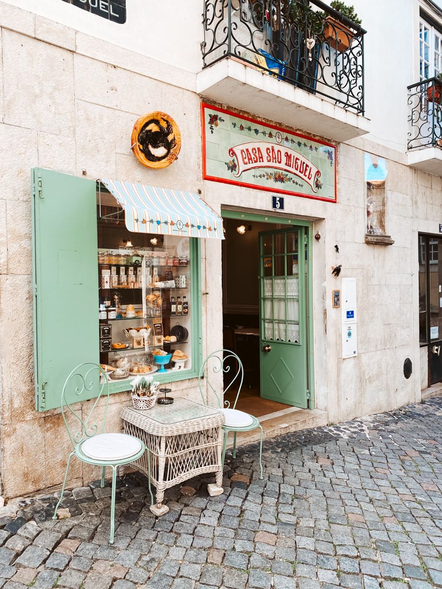 Charming Portuguese shop Casa São Miguel with mint green shutters, outdoor bistro table, and cobblestone street in Lisbon.