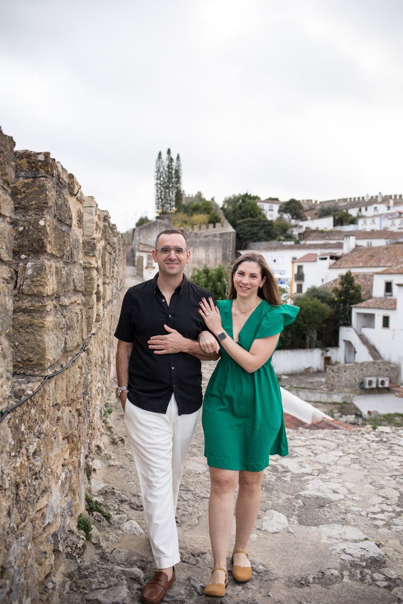 Couple posing on ancient stone wall in European village, woman in green dress showing engagement ring, man in black shirt.