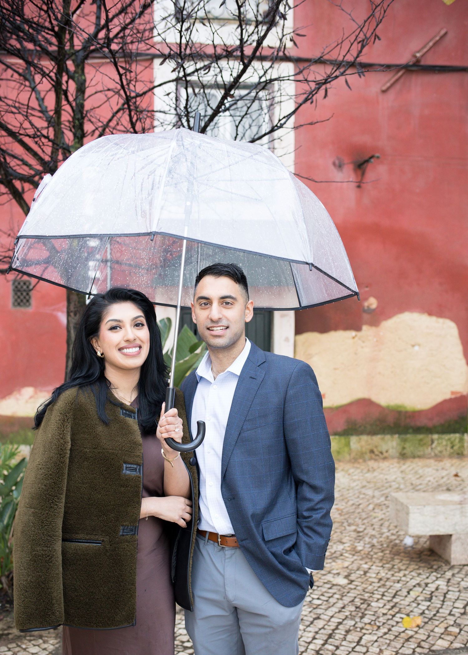 Couple sharing a clear umbrella outdoors, man in blue blazer, woman in olive coat, red wall background.