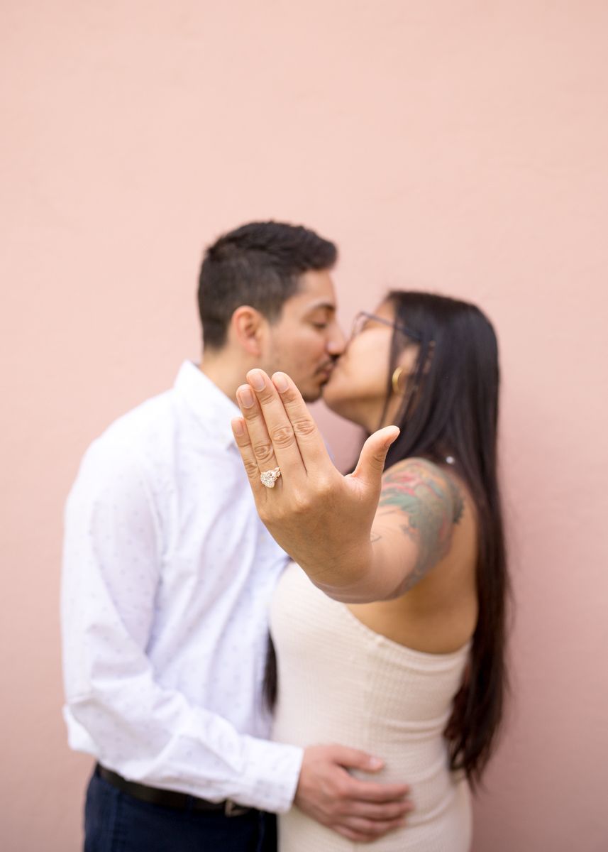Couple sharing a kiss while showing off an engagement ring close to the camera against a pink wall.