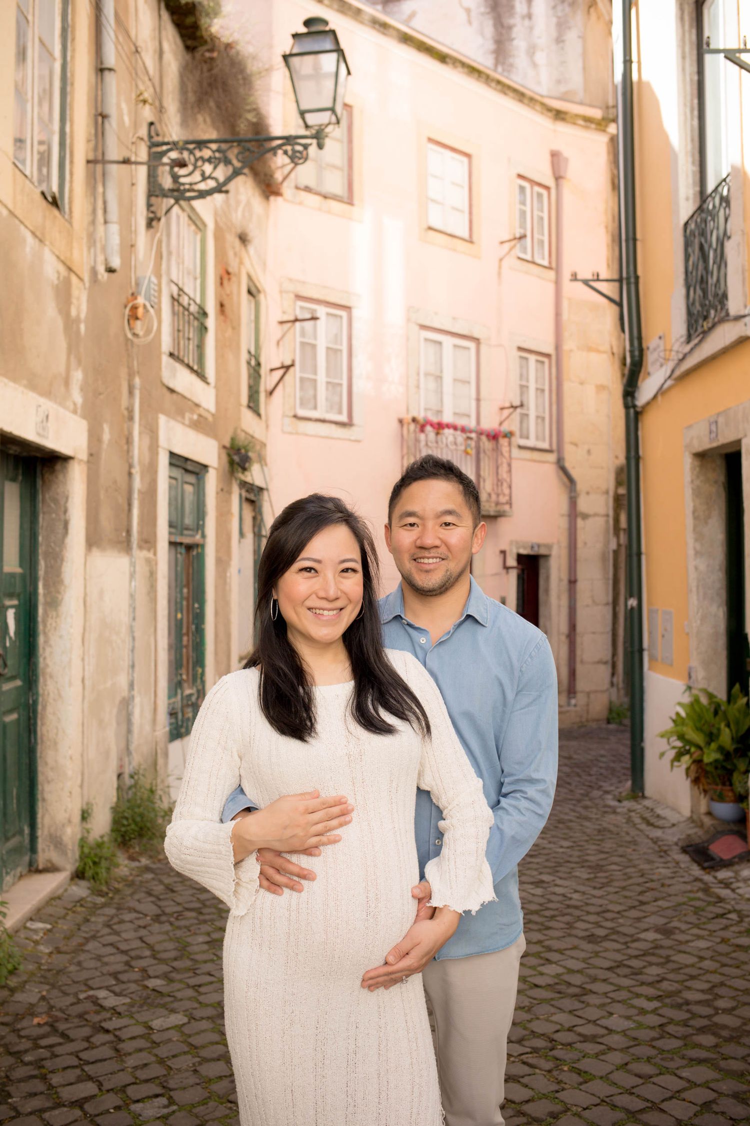 Pregnant woman in white dress and partner embracing on a golden-lit cobblestone alley in Lisbon.