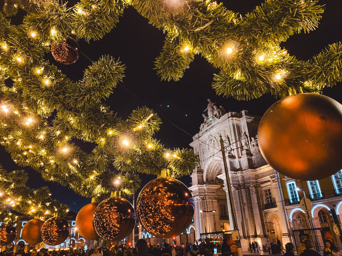 Christmas tree with golden lights and large ornate orange baubles in a festive night square with a historic arch backdrop.