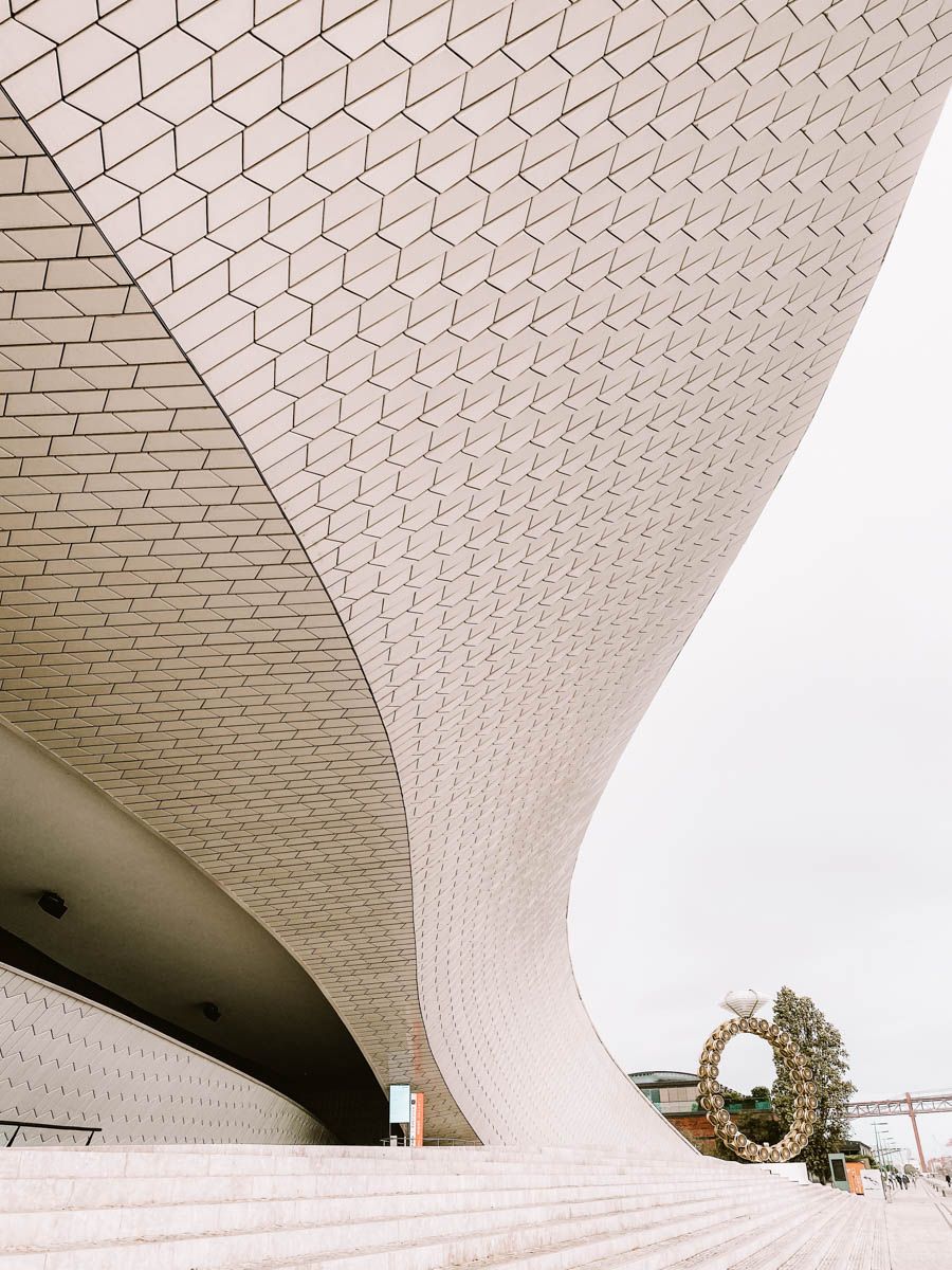 Curved white geometric tile facade of the MAAT museum in Lisbon, Portugal, with steps and overcast sky.
