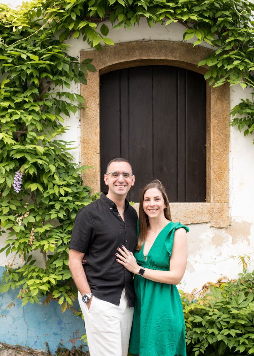 Smiling couple posing in front of a rustic wooden door surrounded by lush green ivy, man in black shirt, woman in green dress.