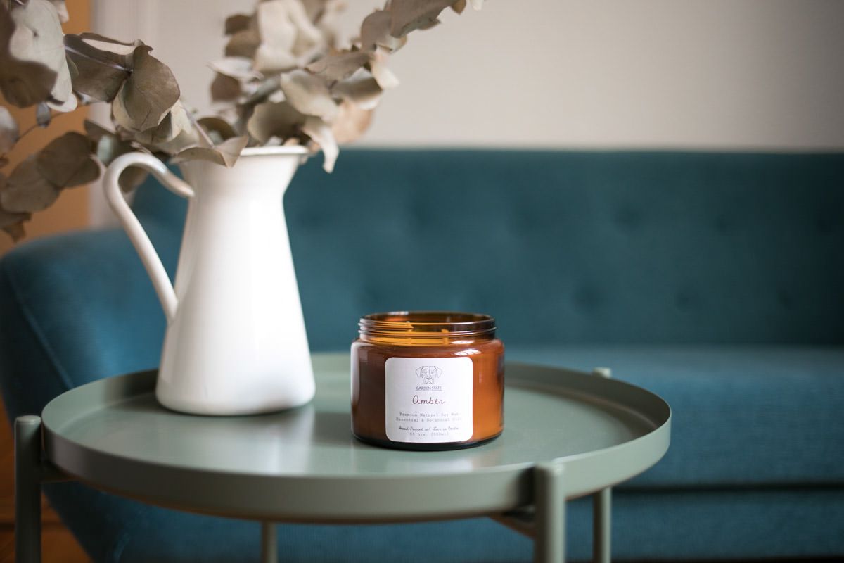 Amber glass candle jar on sage green side table with white ceramic vase and dried eucalyptus, teal sofa background.