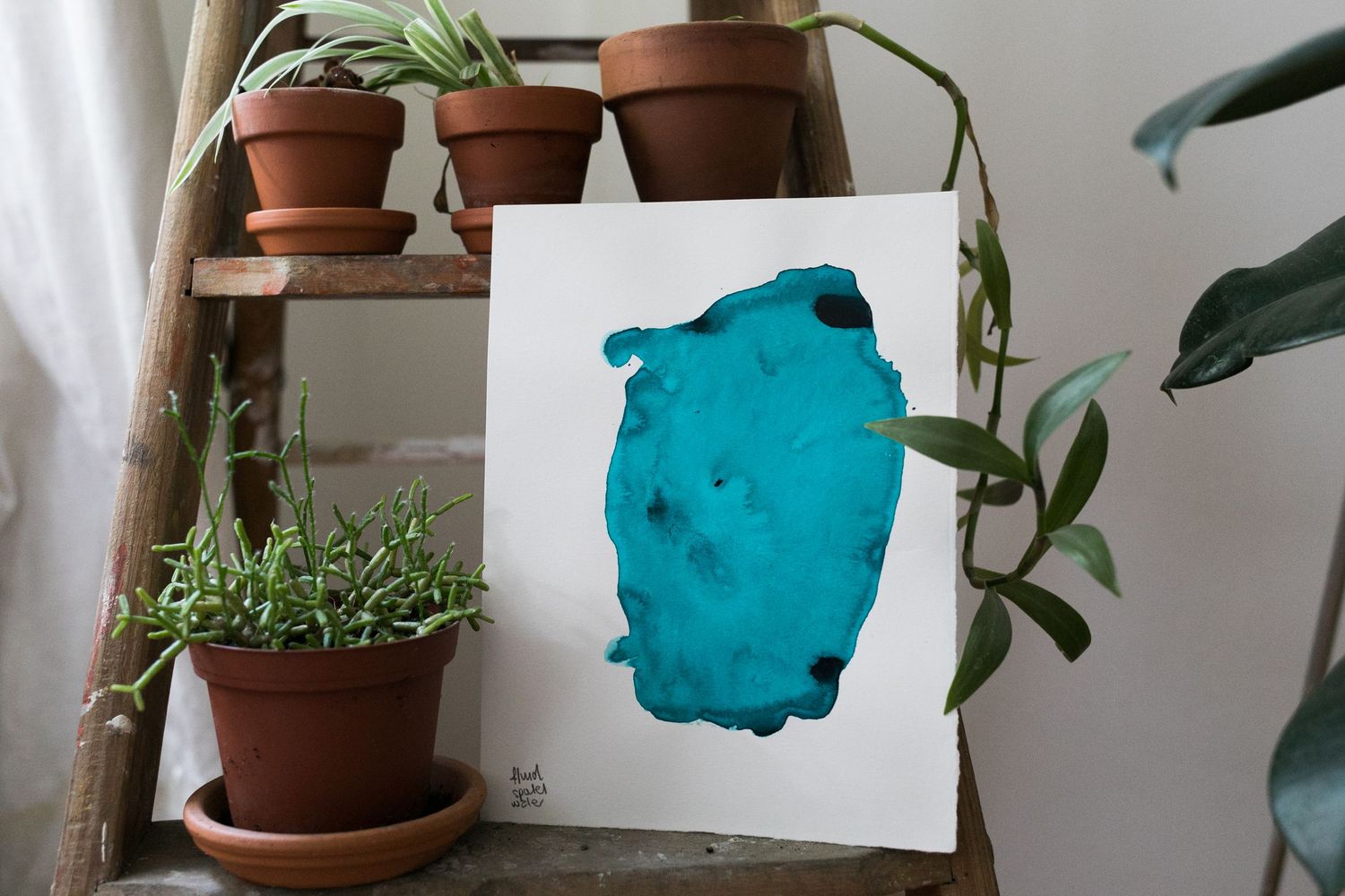 Abstract teal watercolor painting on white paper leaning against a wall, beside potted green plants on a wooden ladder shelf.