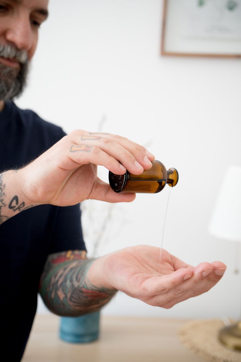 Tattooed bearded man pouring oil from amber bottle into palm indoors.