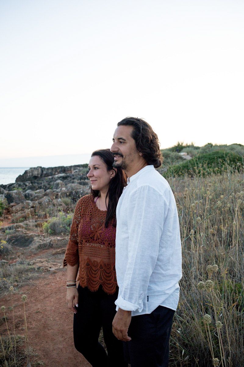 A couple smiles together on a coastal trail at dusk, woman in brown crochet top, man in white linen shirt.