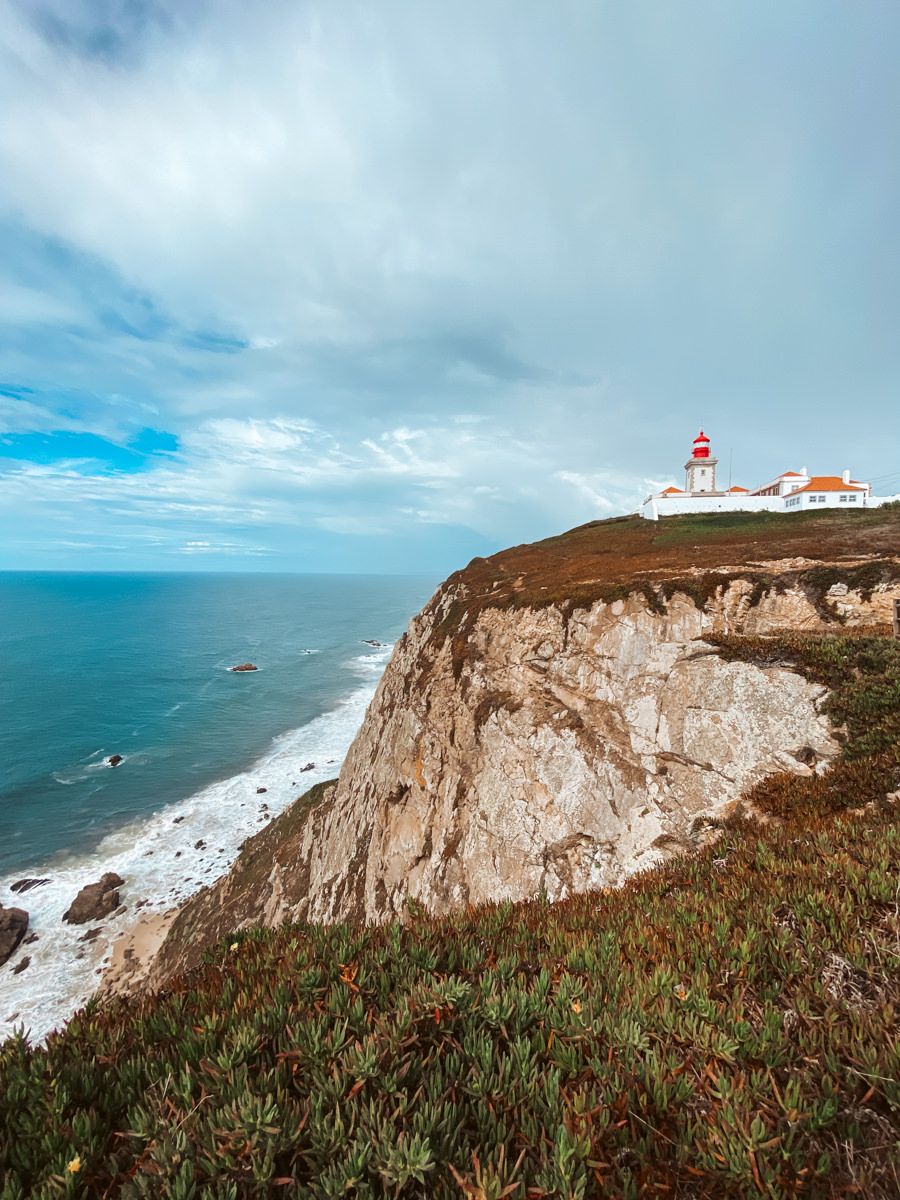 A red and white lighthouse perched atop dramatic rocky cliffs overlooking the Atlantic Ocean under a cloudy sky.