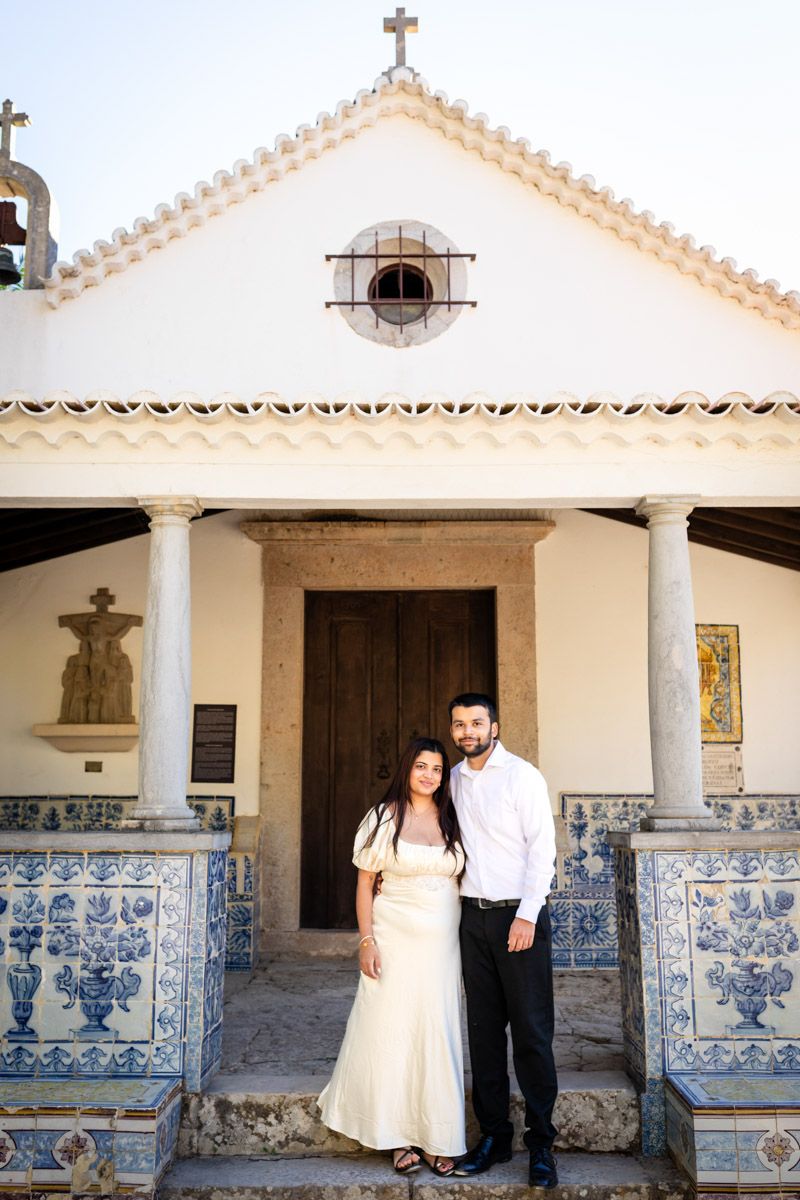 Engaged couple standing together before white chapel adorned with blue Portuguese azulejo tiles.