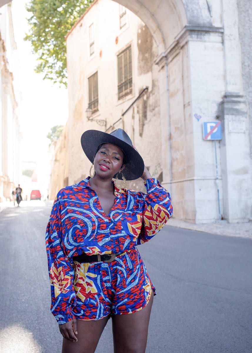 Stylish woman in colorful printed romper and wide-brim hat posing on sunny city street.