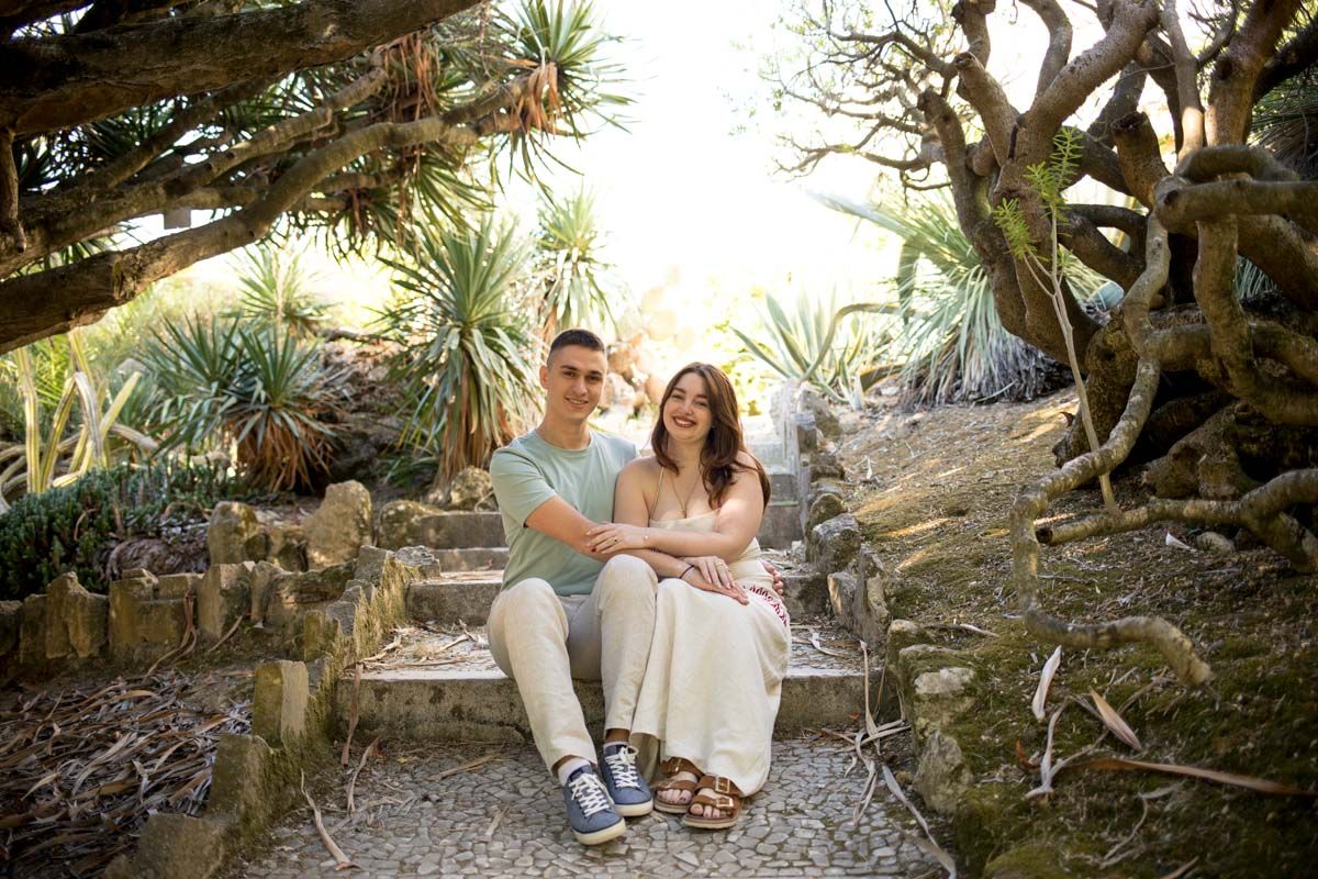 A couple sits together on stone steps surrounded by lush cacti and tropical plants in a scenic garden.
